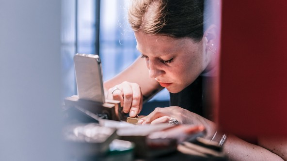 Junge Frau sitzt an Werktisch und arbeitet präzise mit einer Pinzette.