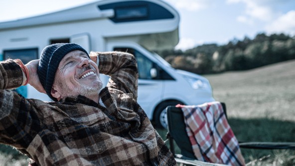 Älterer Mann mit blauer Mütze sitzt lächelnd auf einem Stuhl vor einem Wohnmobil