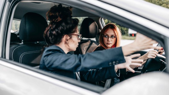 Frau sitzt auf Beifarersitz eines Autos und erklärt einer jungen Frau am Fahrersitz etwas