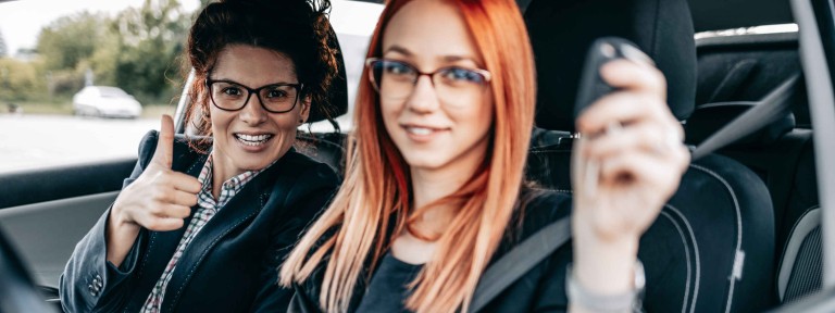 Junge Frau mit Brille sitzt auf Fahrersitz eines Autos und hält den Schlüssel nach oben - am Beifahrersitz blickt eine etwas ältere Frau in die Kamera